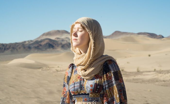 Woman in flowing dress and headscarf standing in Mojave Desert dunes near Tecopa, eyes closed in sunlight