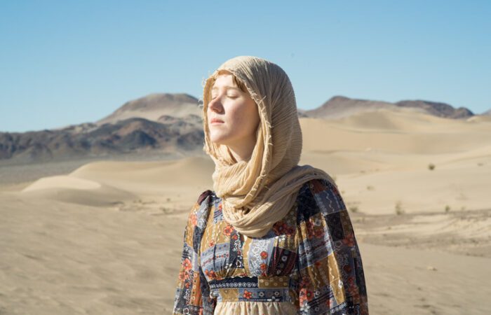 Woman in flowing dress and headscarf standing in Mojave Desert dunes near Tecopa, eyes closed in sunlight