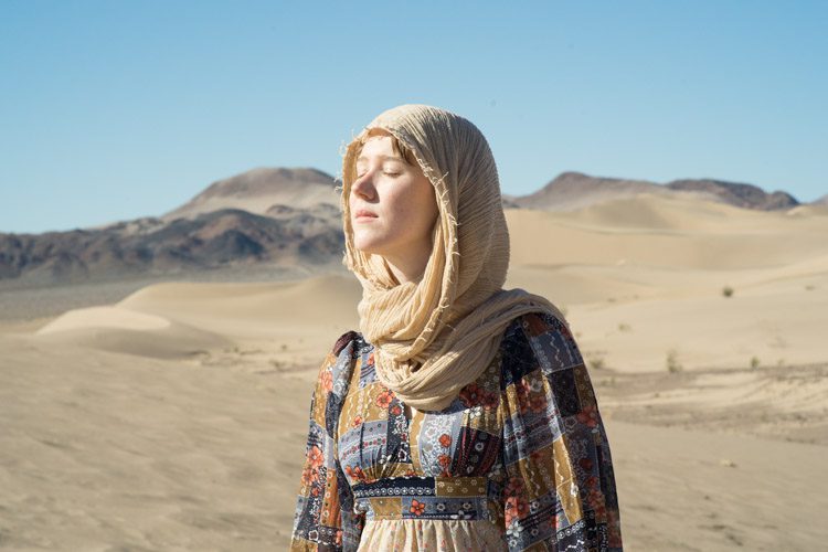Woman in flowing dress and headscarf standing in Mojave Desert dunes near Tecopa, eyes closed in sunlight
