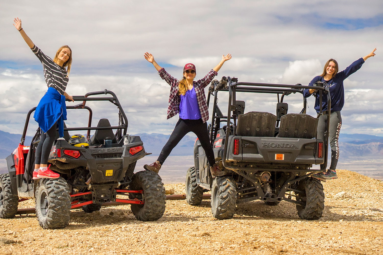 3 women standing on 2 ATVs smiling with arms waving