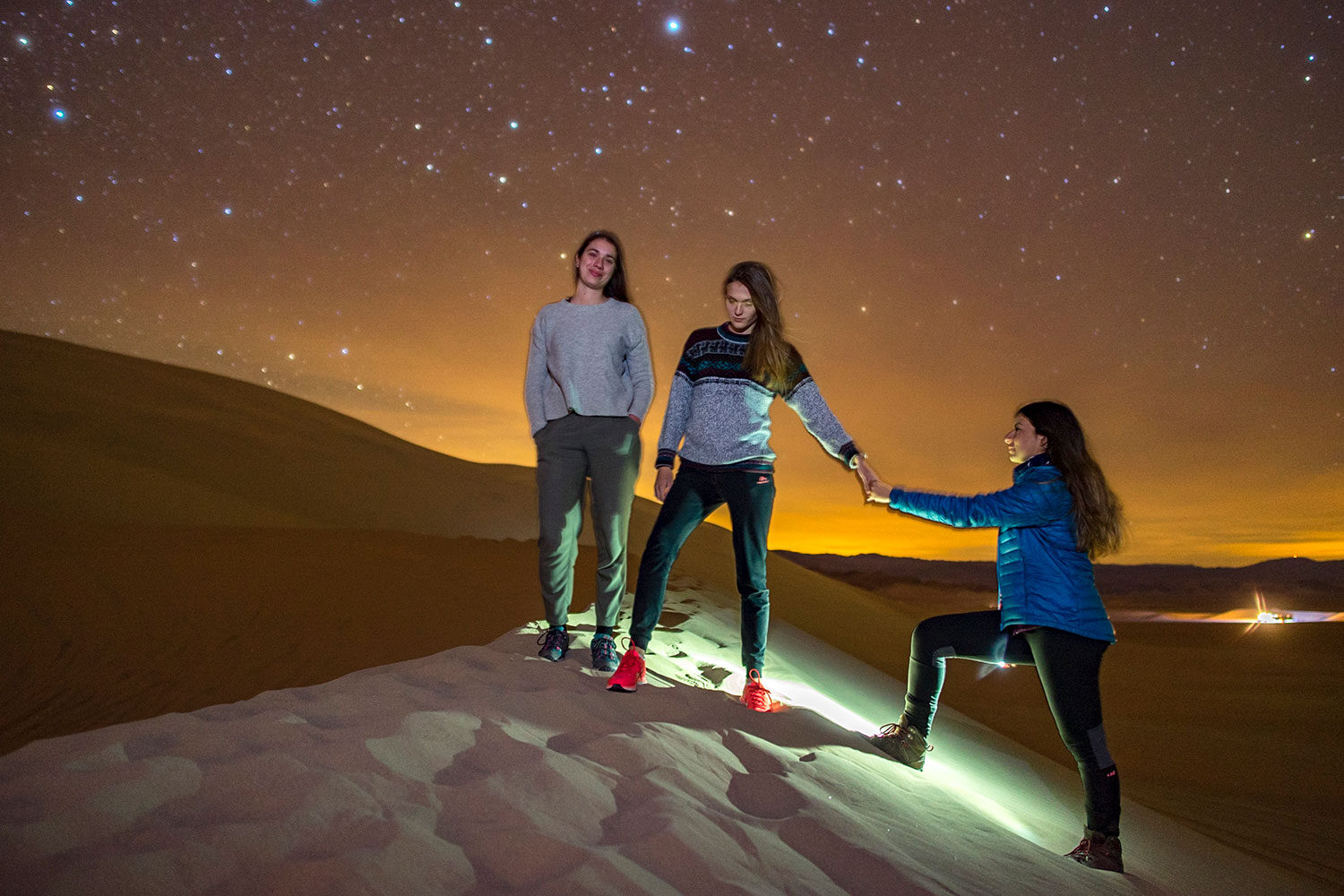 Three women on top of a sand dune under the stars. One women helping to pull another women up.