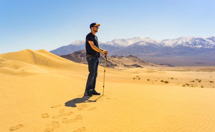 Contemplative man standing on sand dunes surrounded by mountains and blue sky with his hiking pole
