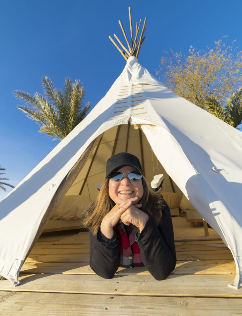 Cynthia smiling in front of a canvas tipi at Cynthia’s Basecamp in Tecopa, California near Death Valley National Park.