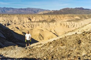 A hiker stands on rocky terrain overlooking arid desert hills under a clear blue sky.